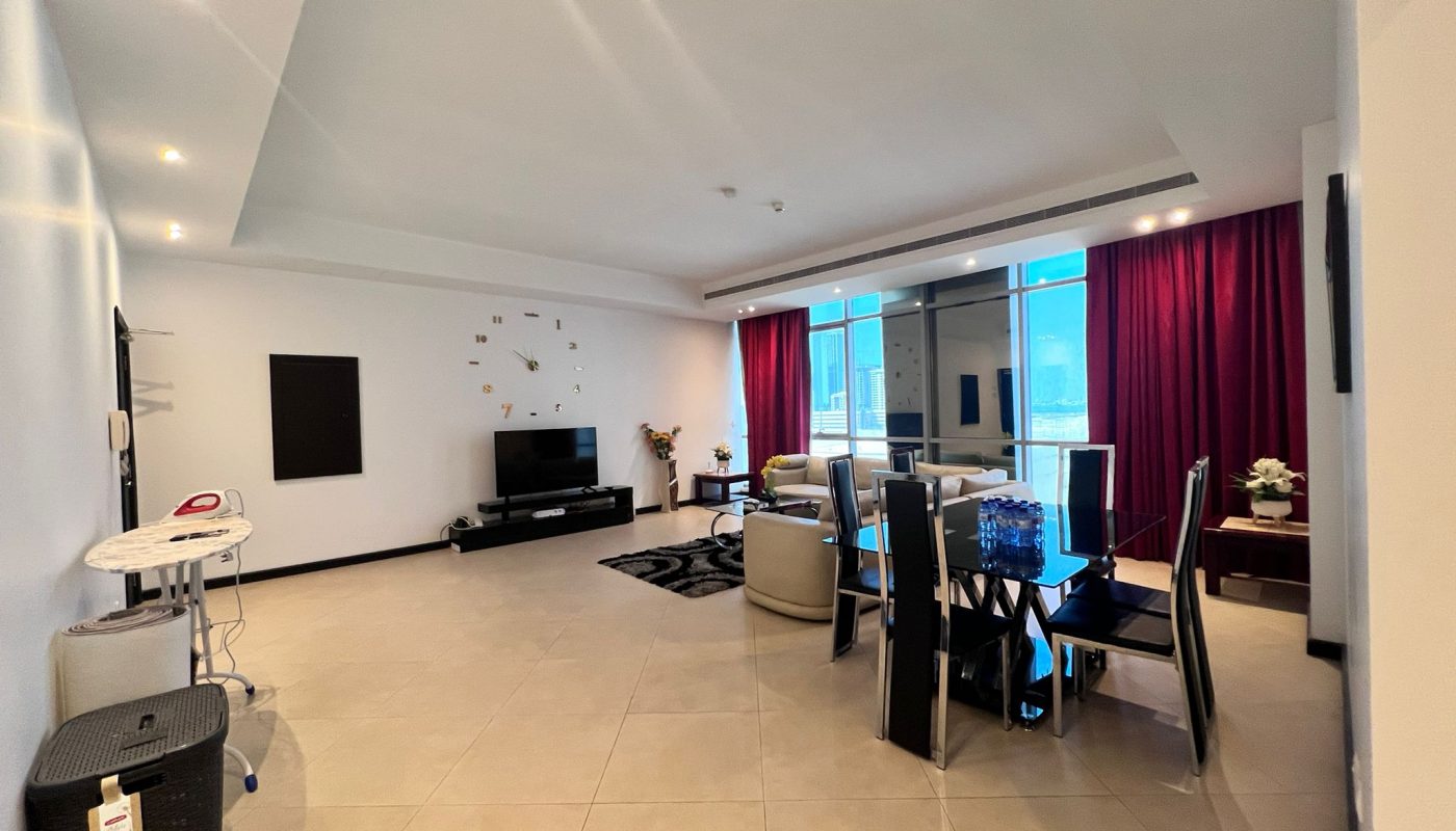 Spacious modern living and dining area with tiled floors, a TV, wall clock, and large windows covered by red curtains.