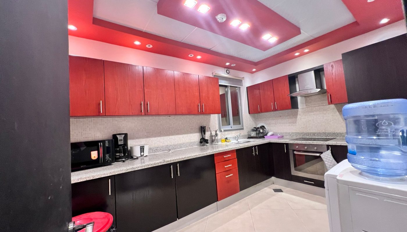 Modern kitchen with red and dark cabinets, stainless steel appliances, a microwave, countertop appliances, and a water dispenser in the corner. Ceiling features red accents and recessed lights.