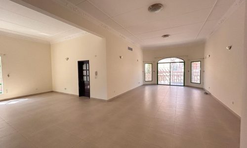 A spacious, empty room with white walls, tiled floor, and large windows. The room has a mix of natural and artificial lighting, with an arched door leading to a balcony.