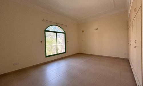 An empty, well-lit room with a large arched window, tiled floor, cream-colored walls, and a built-in wardrobe on one side.