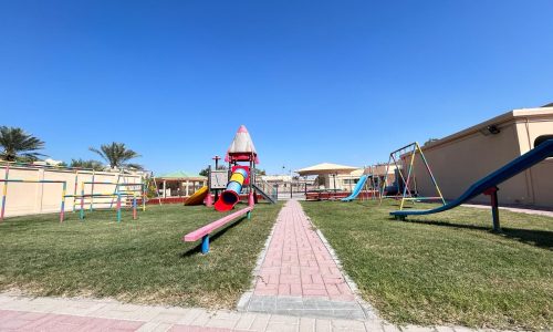 Outdoor playground with various equipment including a slide, swings, and seesaw, on a grassy area surrounded by a paved walkway under a clear blue sky.