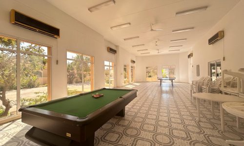 A recreation room featuring a pool table with green felt, stacked white chairs, large windows, wall-mounted air conditioners, and modern patterned floor tiles.