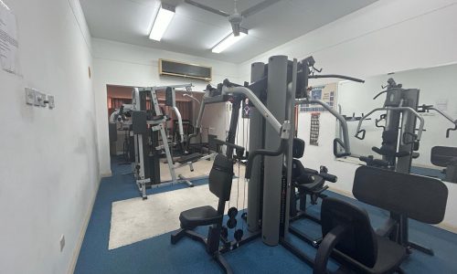 A small gym room with various exercise equipment, including weight machines and benches, a ceiling fan, and fluorescent lighting. The walls are white, and the floor is blue carpet.