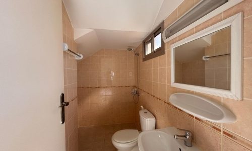 Small bathroom with beige tiled walls, featuring a toilet, sink, mirror, and overhead light. A narrow window is placed above the toilet, and the door is partially open.