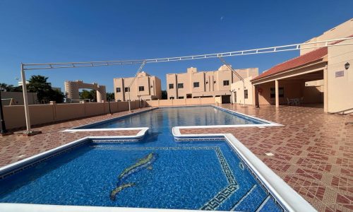 A residential swimming pool area with a main pool and a smaller shallow section. The tiled pool deck has scattered furniture, and rows of houses are seen in the background under a clear blue sky.