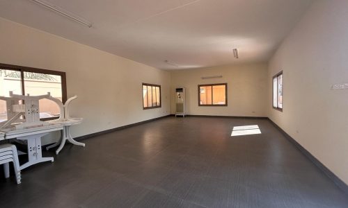 A spacious, empty room with tiled flooring, multiple windows, fluorescent lights, and stacked tables and chairs in one corner.