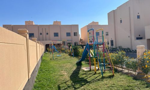 A small grassy playground with colorful climbing frames, slides, and swings is situated between beige residential buildings under a clear blue sky.