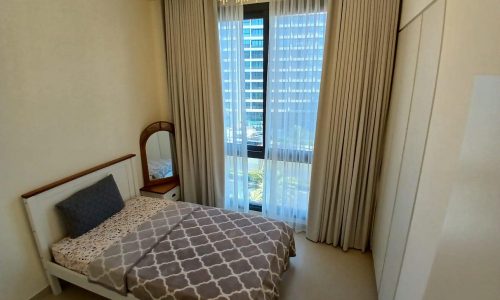 Modern bedroom in a luxury apartment, with a single bed, patterned bedding, floor-to-ceiling windows with sheer curtains, and a chandelier.