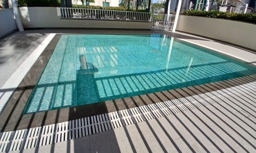 Outdoor swimming pool with clear blue water, surrounded by a tiled deck and shaded by a modern pergola, adjacent to a luxury apartment.