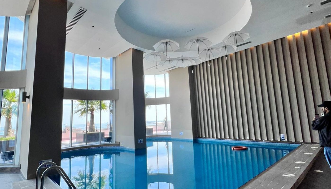 Indoor swimming pool with large windows, high ceiling, and modern design. A person stands near the edge, and palm trees are visible outside through the glass.