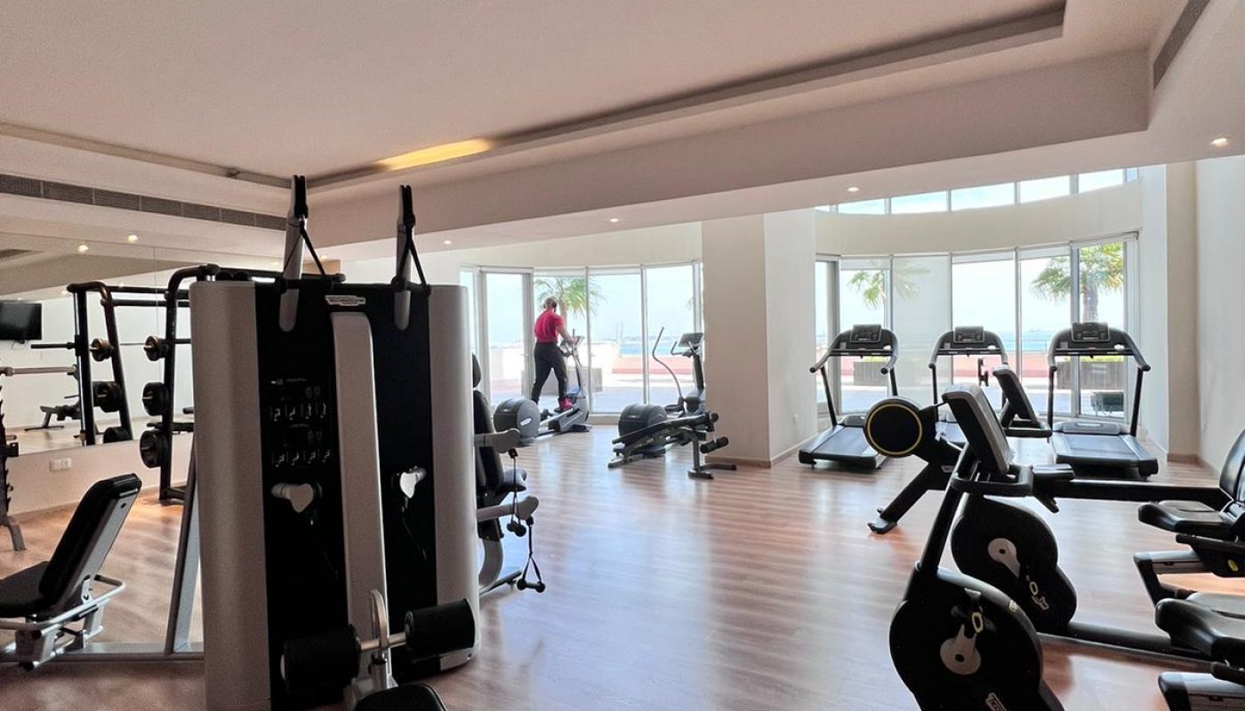 Modern gym with various exercise machines, free weights, and a person using equipment near large windows letting in natural light.