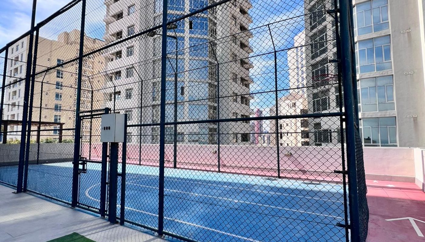 A fenced outdoor sports court with blue and pink surfaces is surrounded by tall modern apartment buildings under a partly cloudy sky.
