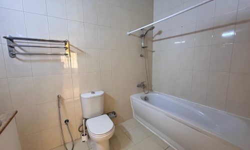 A bathroom with white tiles, featuring a toilet, a bidet sprayer, a bathtub with a shower overhead, and a towel rack.