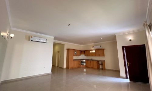 A spacious, unfurnished room with a tiled floor, featuring an open kitchen with wooden cabinets, two air conditioning units, a closed door, and light fixtures on the walls.