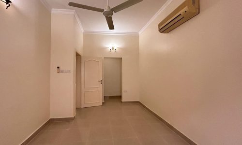 Empty room with tiled floor, beige walls, ceiling fan, air conditioner, and two wall-mounted light fixtures. Door visible in the back leads to another area.