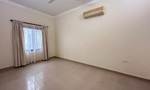 A spacious, empty room with beige walls, a ceiling fan, an air conditioner, a window with curtains, and a tiled floor.