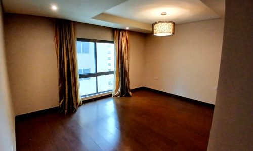 An empty room with wooden flooring, beige walls, a window with auto draft curtains, and a single ceiling light fixture.