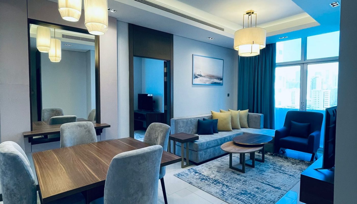 Modern living and dining area with grey chairs, wooden table, blue accent sofa, large windows, pendant lights, and a wall mirror.