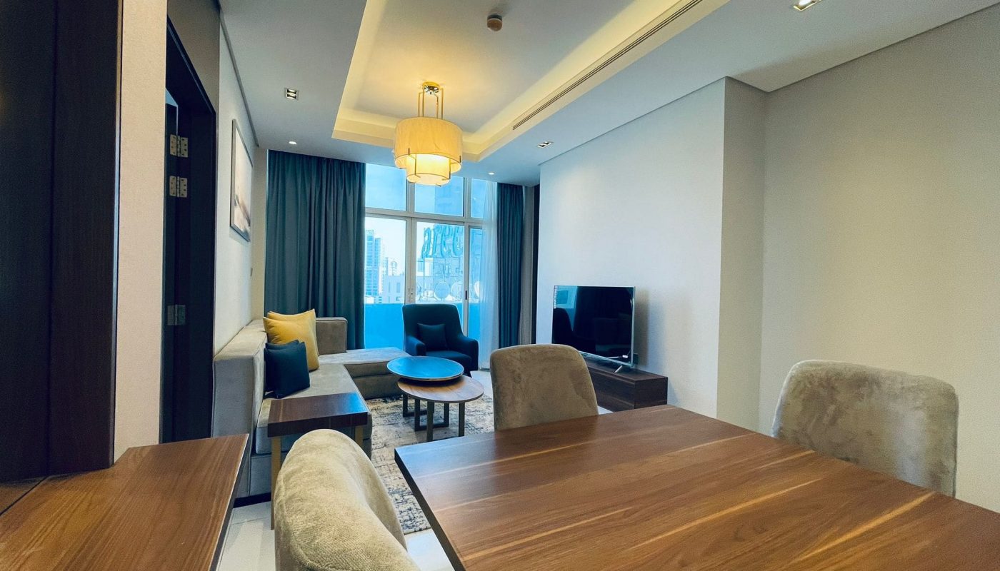 Modern living room with dining area in a 1BR apartment, featuring an eye-catching wooden table, plush seating, and a television.