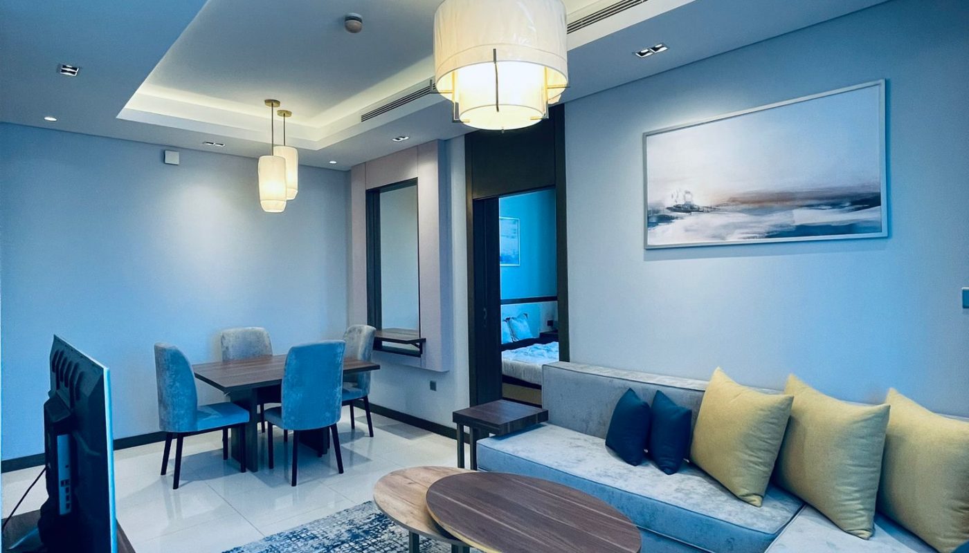Modern living room with a blue and beige color scheme, sofa, armchair, coffee tables, TV, dining table, and wall art under ceiling lights.