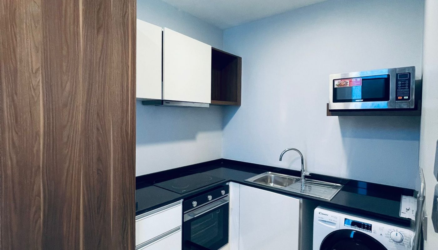Modern kitchen with white cabinets, black countertops, built-in oven, sink, washing machine, and a microwave on the counter under bright ceiling lights.