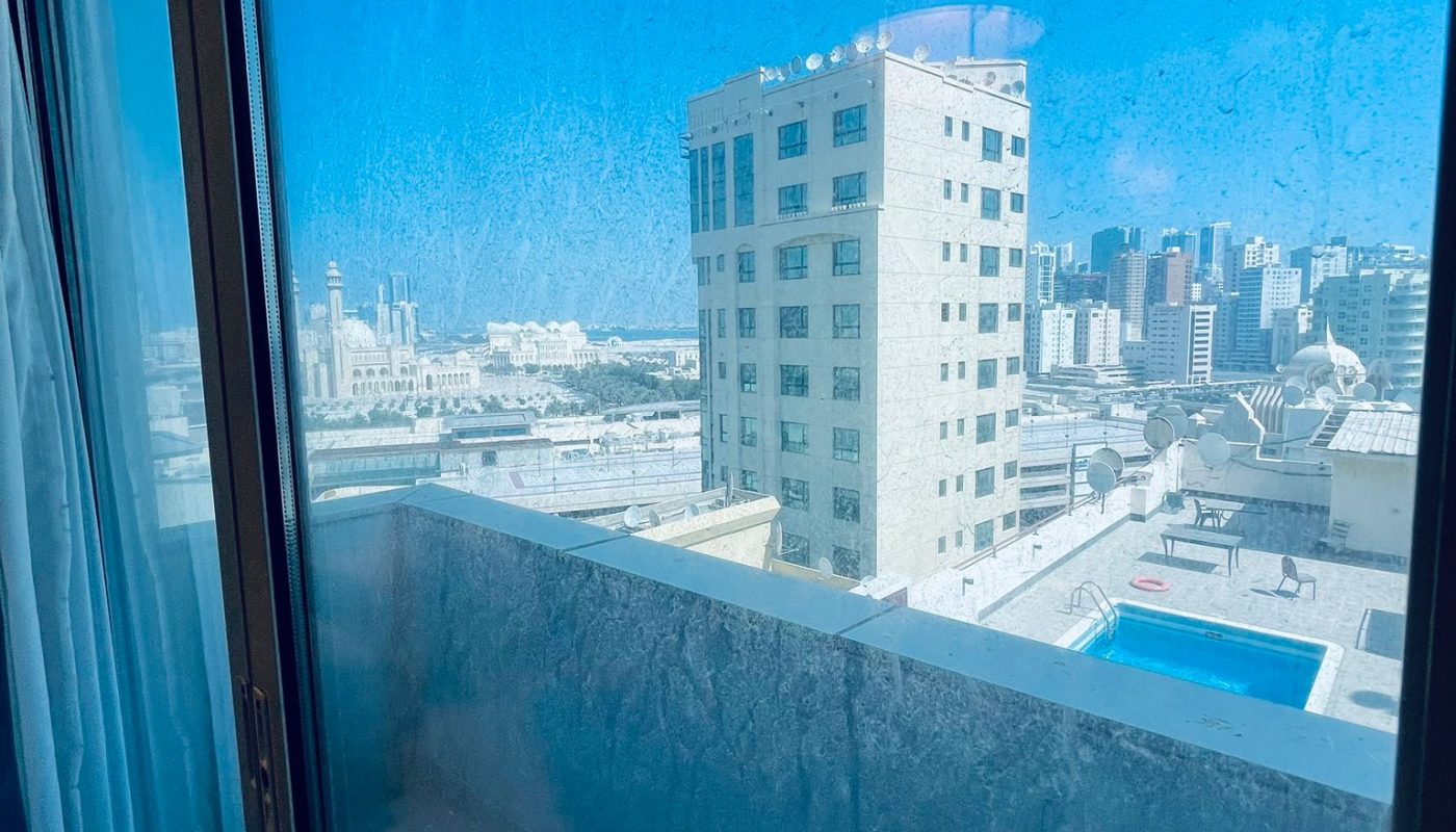 View through a glass door showing a small balcony, a tall building, a pool, and a cityscape under a clear blue sky.