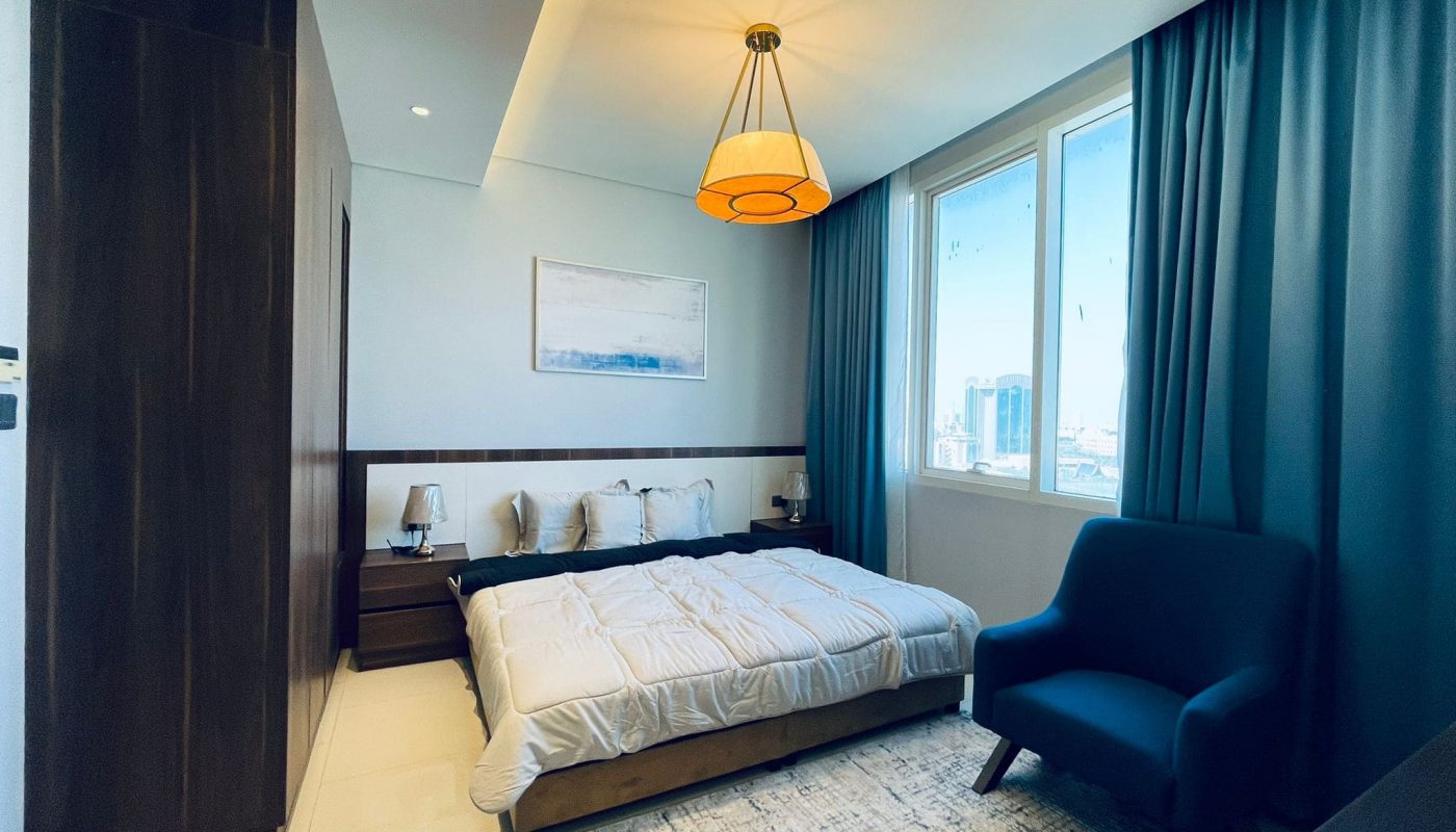 Modern 1BR apartment bedroom in Juffair with a large window offering an eye-catching city view, featuring a neatly made bed, blue armchair, and an overhead pendant light.