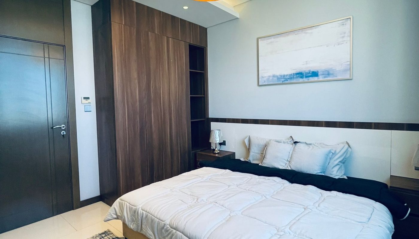 Modern bedroom with a double bed, white bedding, wooden wardrobe, abstract wall art, and a ceiling light fixture. A patterned rug and dark blue chair are also visible.