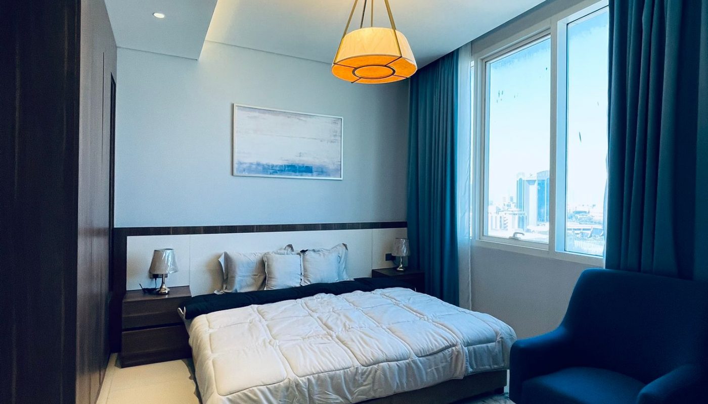Modern bedroom with a large bed, blue curtains, accent chair, nightstands, a ceiling light, and a city view through a window.