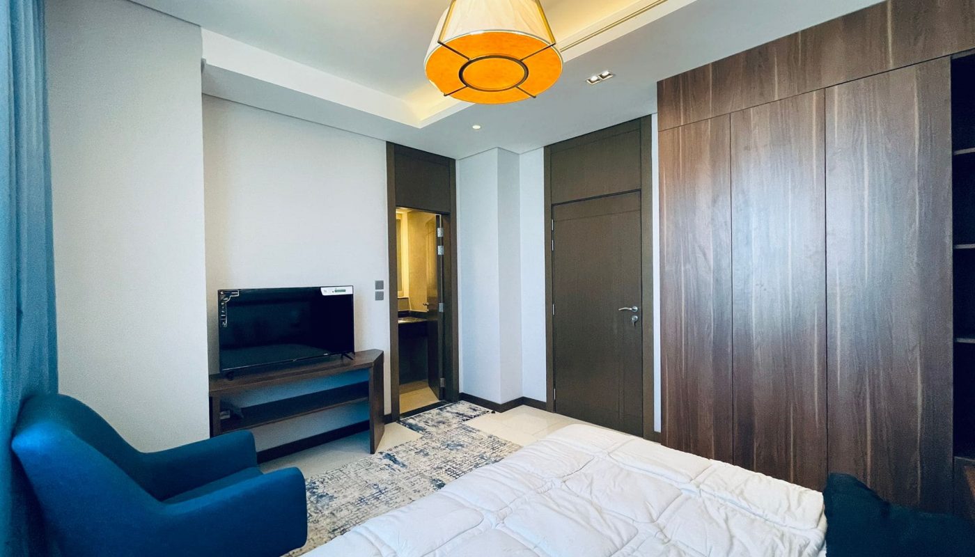 Eye-catching 1BR apartment in Juffair with a neatly made bed, a blue armchair, wooden wardrobe, and a flat-screen TV.