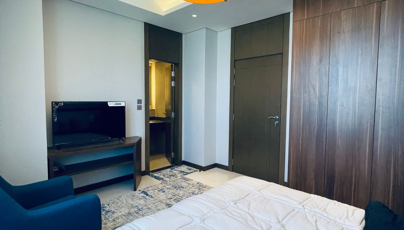 Modern bedroom with a double bed, blue armchair, TV on a stand, wooden wardrobe, and a ceiling light fixture. Doorway leads to another room.