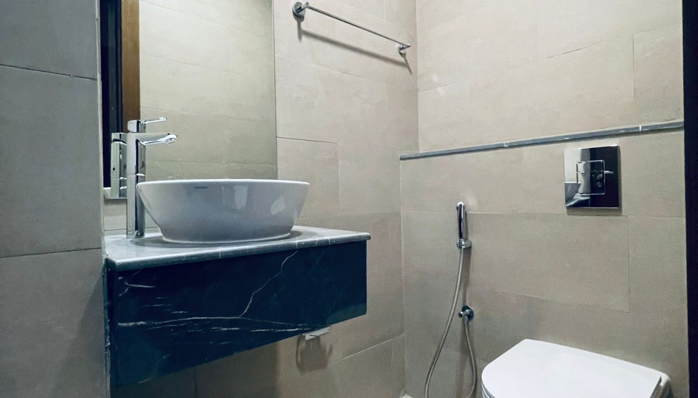 Modern bathroom with a wall-mounted toilet, vessel sink on a marble counter, large mirror, towel rack, and handheld bidet sprayer on tiled walls and floor.