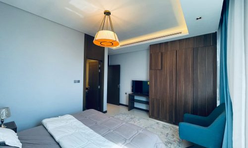 Modern bedroom interior in a Juffair apartment for rent with a large bed, wooden wardrobe, blue armchair, and pendant light.