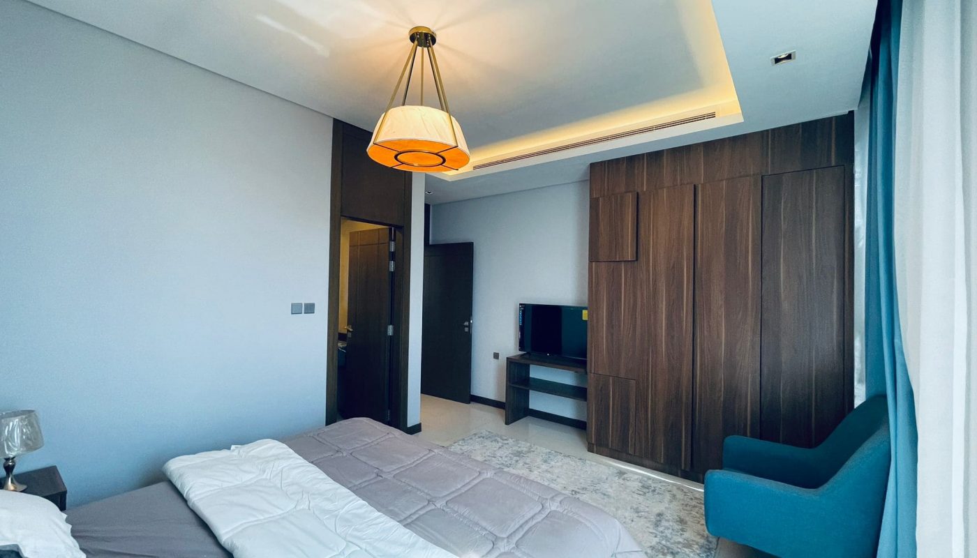 Modern bedroom interior in a Juffair apartment for rent with a large bed, wooden wardrobe, blue armchair, and pendant light.