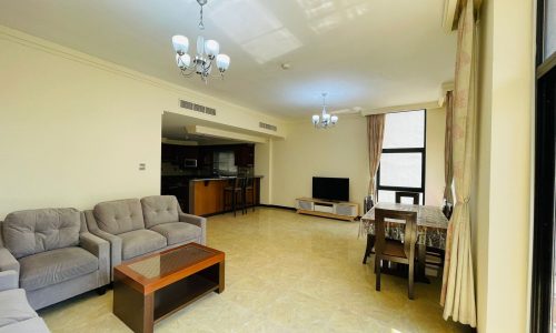 A well-lit, spacious living room with a sofa, a coffee table, and an open-plan kitchen in the background. This space is part of an apartment for rent in Juffair.