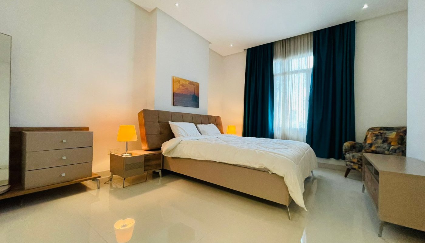 A well-lit modern bedroom featuring a double bed with a beige headboard, white bedding, two bedside tables with lamps, an armchair, all complemented by dark blue curtains and a decorative painting