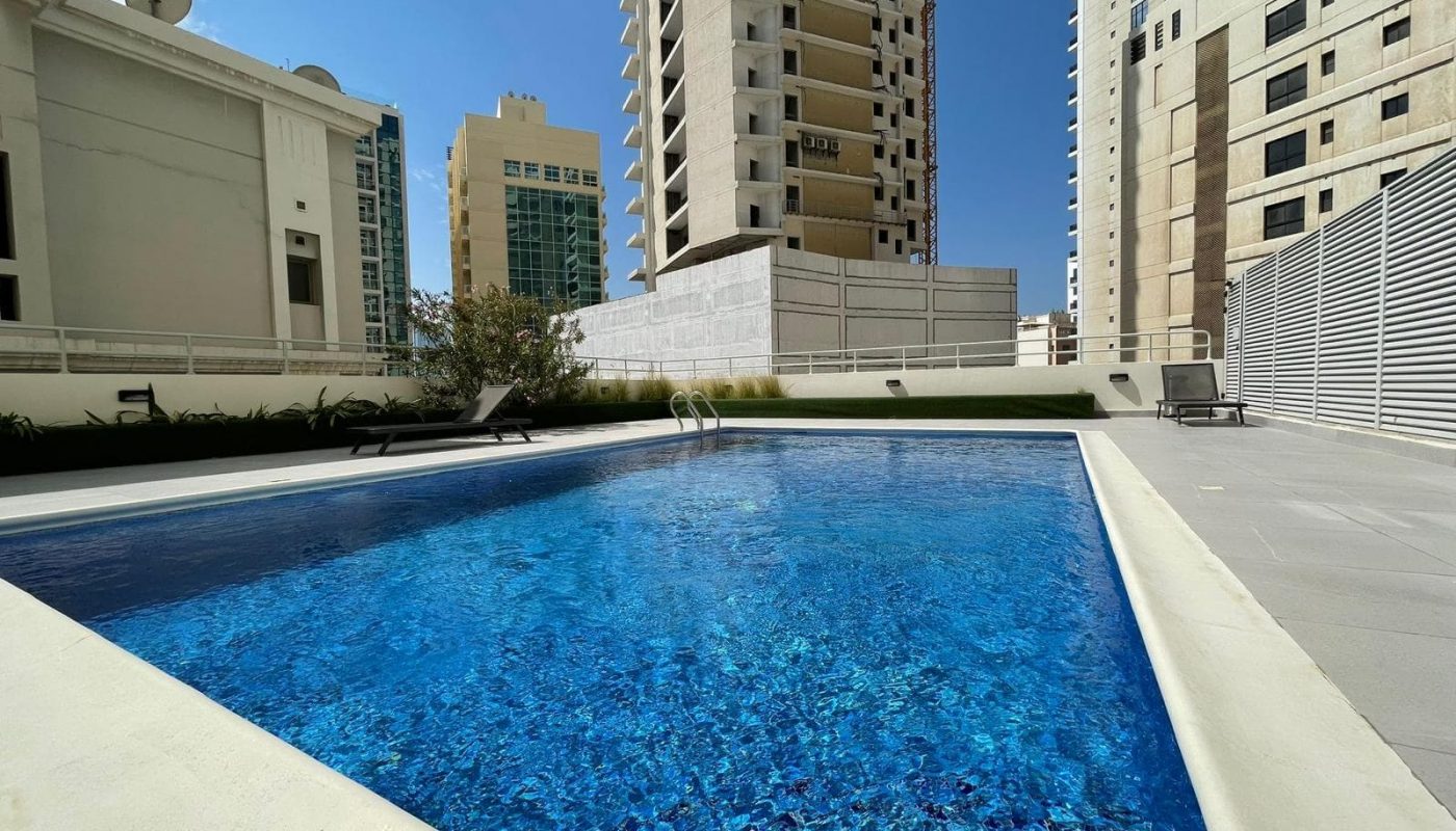 An outdoor swimming pool with clear blue water in an urban environment surrounded by tall buildings and auto drafts.