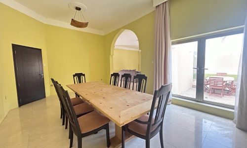 Spacious dining room in a Hamala area villa for rent, featuring a large wooden table, green walls, and access to an outdoor patio area.