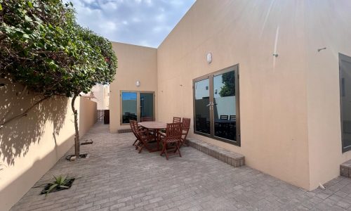 A paved outdoor patio area at the Compound Hamala, with a wooden table and chairs set, surrounded by beige walls and green shrubbery under a clear sky.