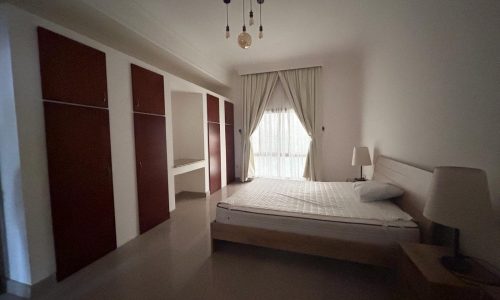 A minimalist bedroom in a villa for rent, featuring a large bed, two-tone wardrobe, side table with a lamp, and sheer curtains on a window in the Budaiya area.