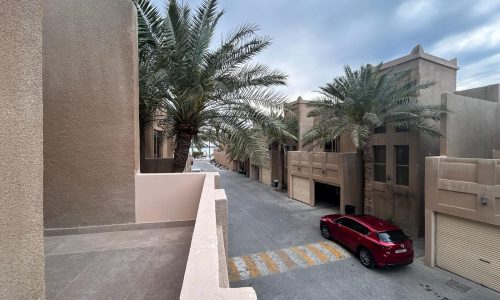 A quiet residential street in the Budaiya area lined with palm trees and villas for rent, featuring a red car parked on the side.