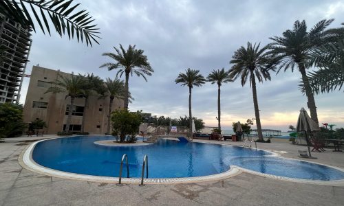 Curved outdoor swimming pool surrounded by palm trees in the Compound Budaiya area, under an overcast sky.