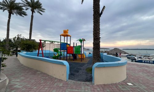 A colorful children's playground by the waterfront in the Compound Budaiya area, with palm trees and an overcast sky.