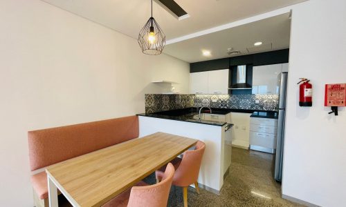 A luxury 1 BR apartment with a modern kitchen and dining area, featuring a wooden table, pink chairs, pendant lighting, and a fire extinguisher on the wall.