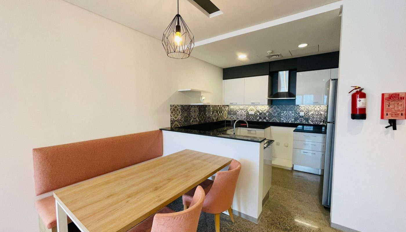 A luxury 1 BR apartment with a modern kitchen and dining area, featuring a wooden table, pink chairs, pendant lighting, and a fire extinguisher on the wall.