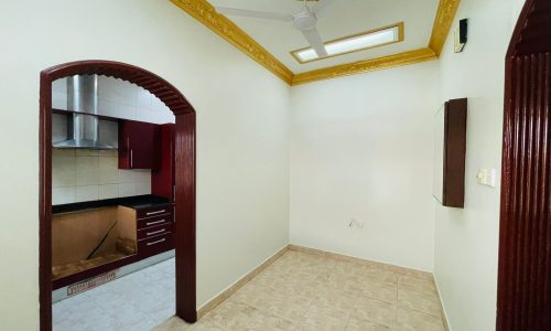 A semi-furnished 2BR villa in Ras Rumman, with tiled flooring featuring an arched entrance to a kitchen with wooden cabinetry and a ceiling fan.