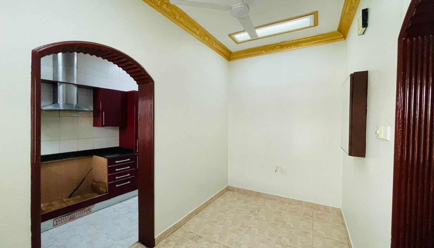 A semi-furnished 2BR villa in Ras Rumman, with tiled flooring featuring an arched entrance to a kitchen with wooden cabinetry and a ceiling fan.