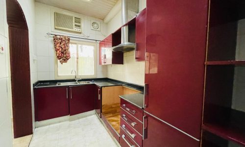 A compact kitchen in a 2BR Villa with maroon cabinets, white tiled flooring, and an air conditioning unit above the window.