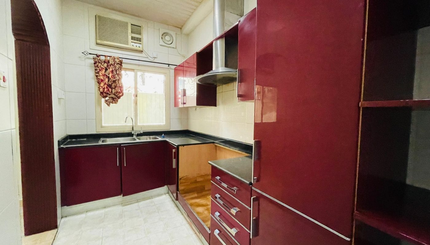 A compact kitchen in a 2BR Villa with maroon cabinets, white tiled flooring, and an air conditioning unit above the window.