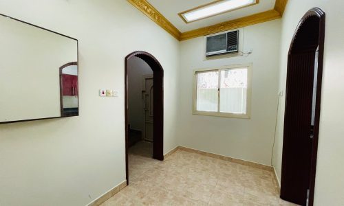 An empty room in a 2BR Villa located in Ras Rumman, featuring a tiled floor, white walls, an air conditioning unit, a window with decorative grills, and two doorways.